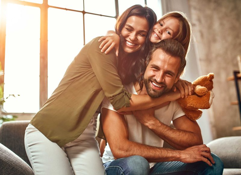 happy-dad-and-mom-with-their-cute-daughter-and-teddy-bear-hug-and-have-fun-sitting-on-the-sofa-in-the-living-room-at-home